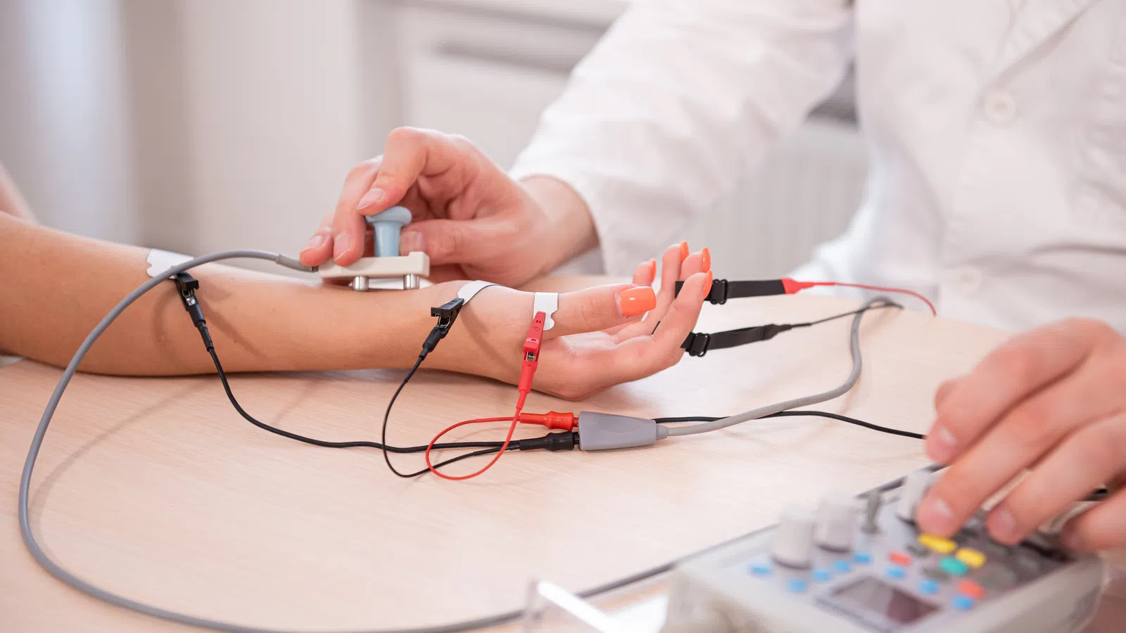 Doctor performing nerve conduction study on patient's hand with electrodes and medical device on a wooden table.