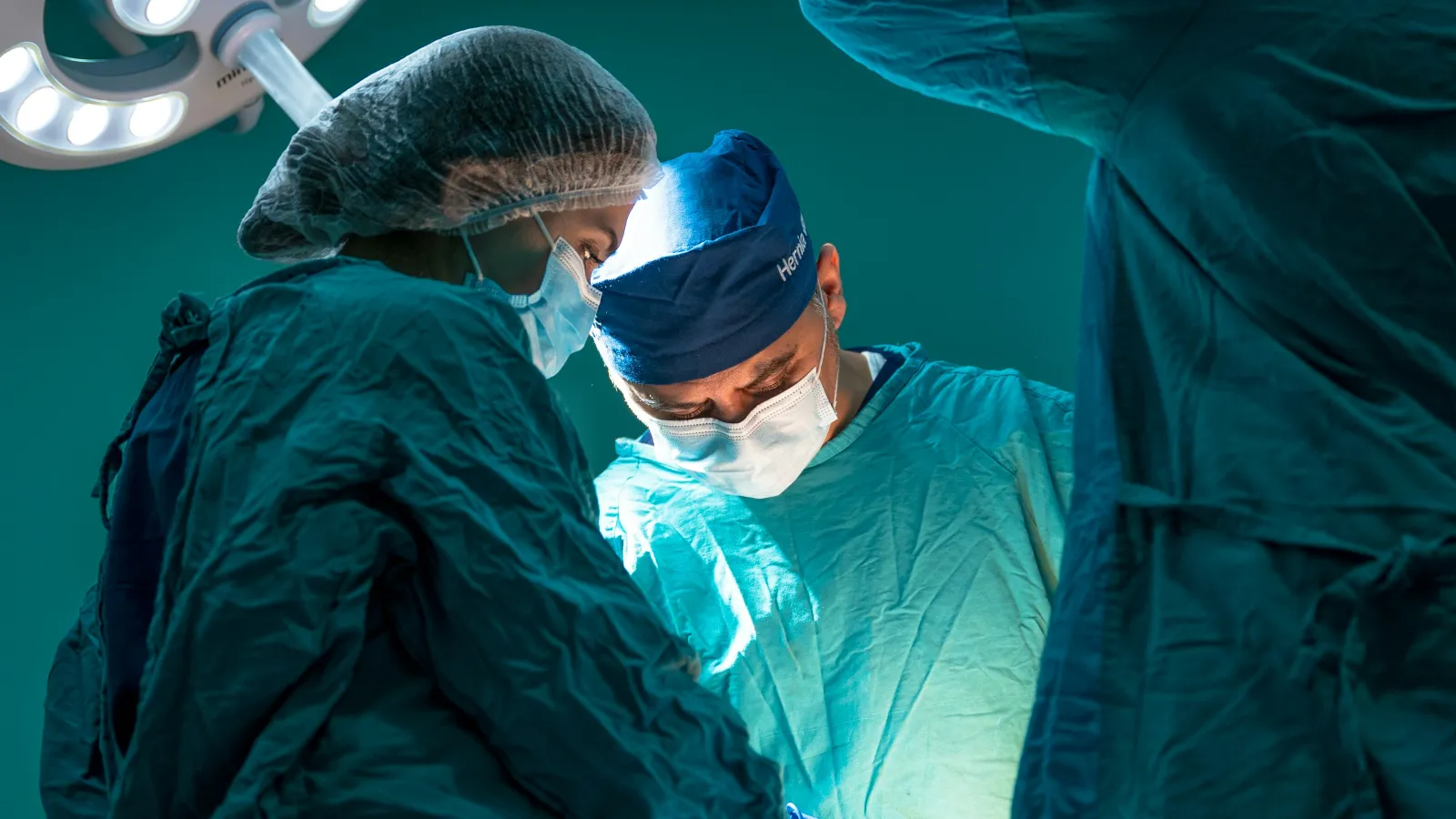 Surgeons in blue scrubs perform surgery under bright operating room lights in a sterile environment