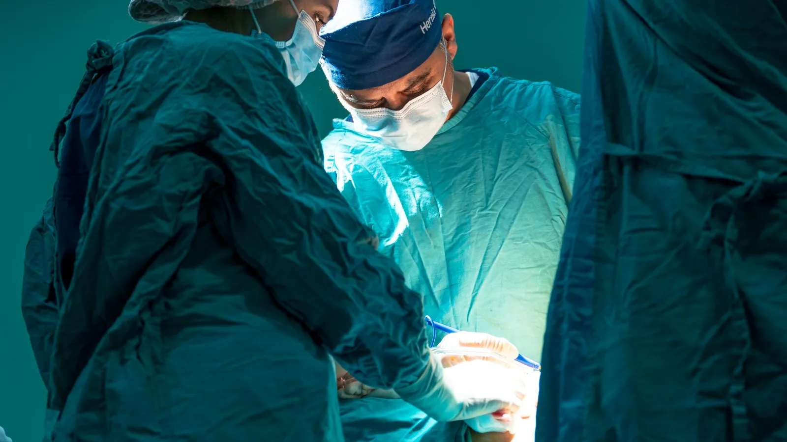 Surgeons in blue scrubs perform surgery under bright operating room lights in a sterile environment