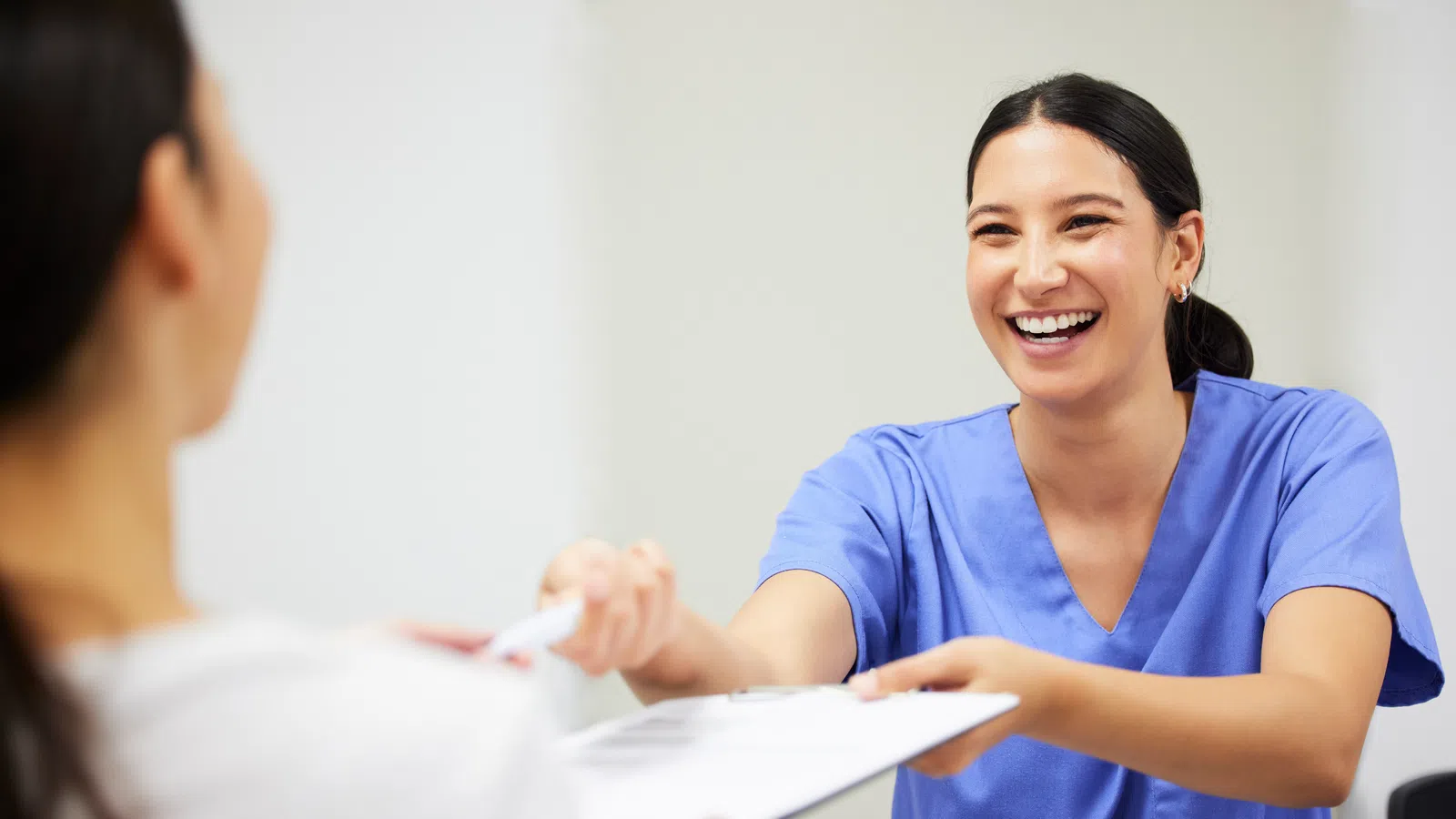 Smiling healthcare professional in blue scrubs handing a clipboard to a patient in a medical setting.