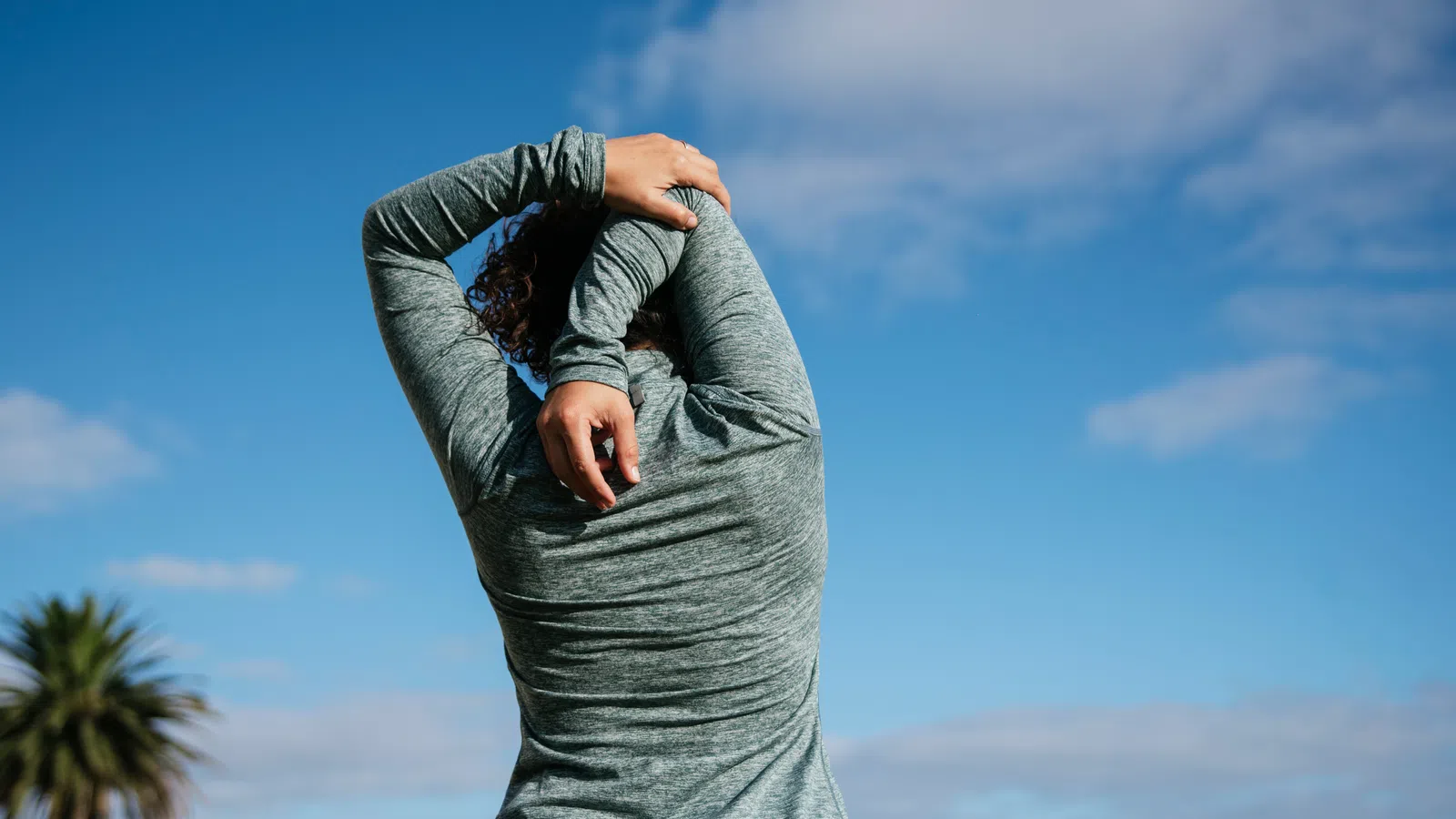 Person in gray hoodie stretching arms overhead outdoors with blue sky and palm tree in background