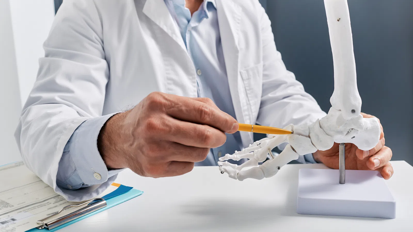 Doctor in white coat pointing at anatomical foot skeleton model with a pencil on white desk.