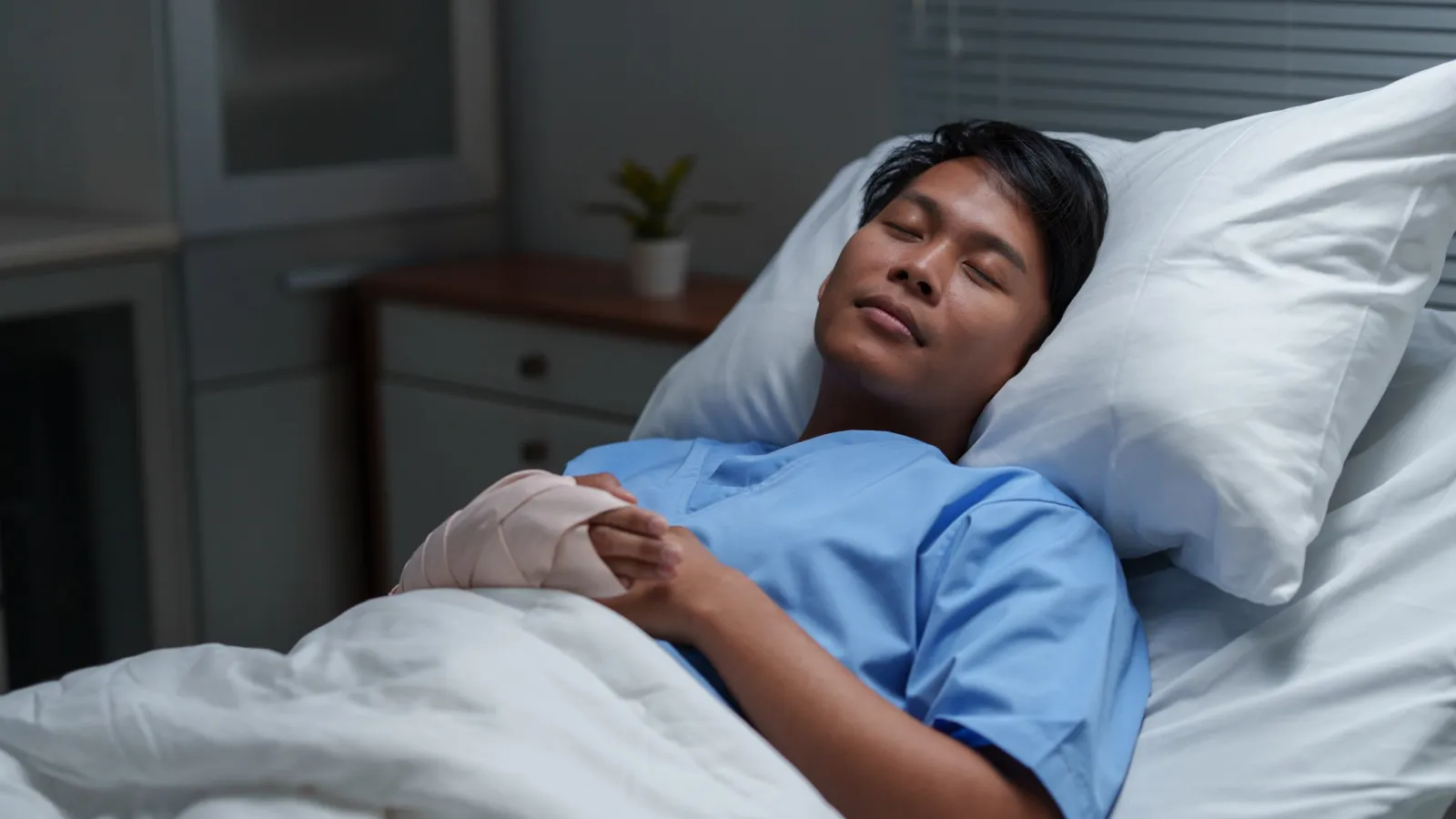 Patient in hospital bed resting with a bandaged hand, wearing a blue medical gown, eyes closed peacefully.