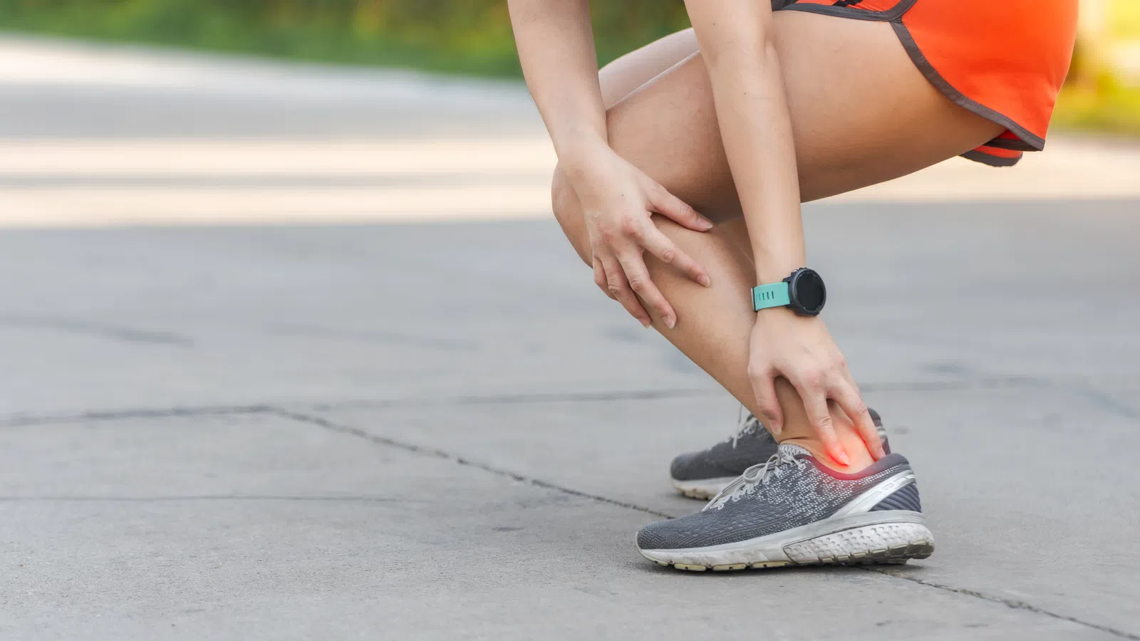 Runner wearing orange shorts clutching ankle in pain on concrete road, highlighting ankle injury area in red.