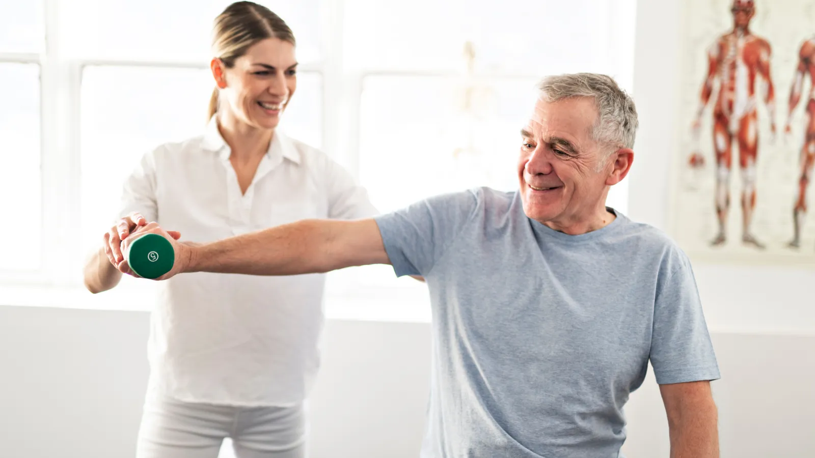 Senior man exercising with a dumbbell assisted by a smiling female physiotherapist in a bright clinic.