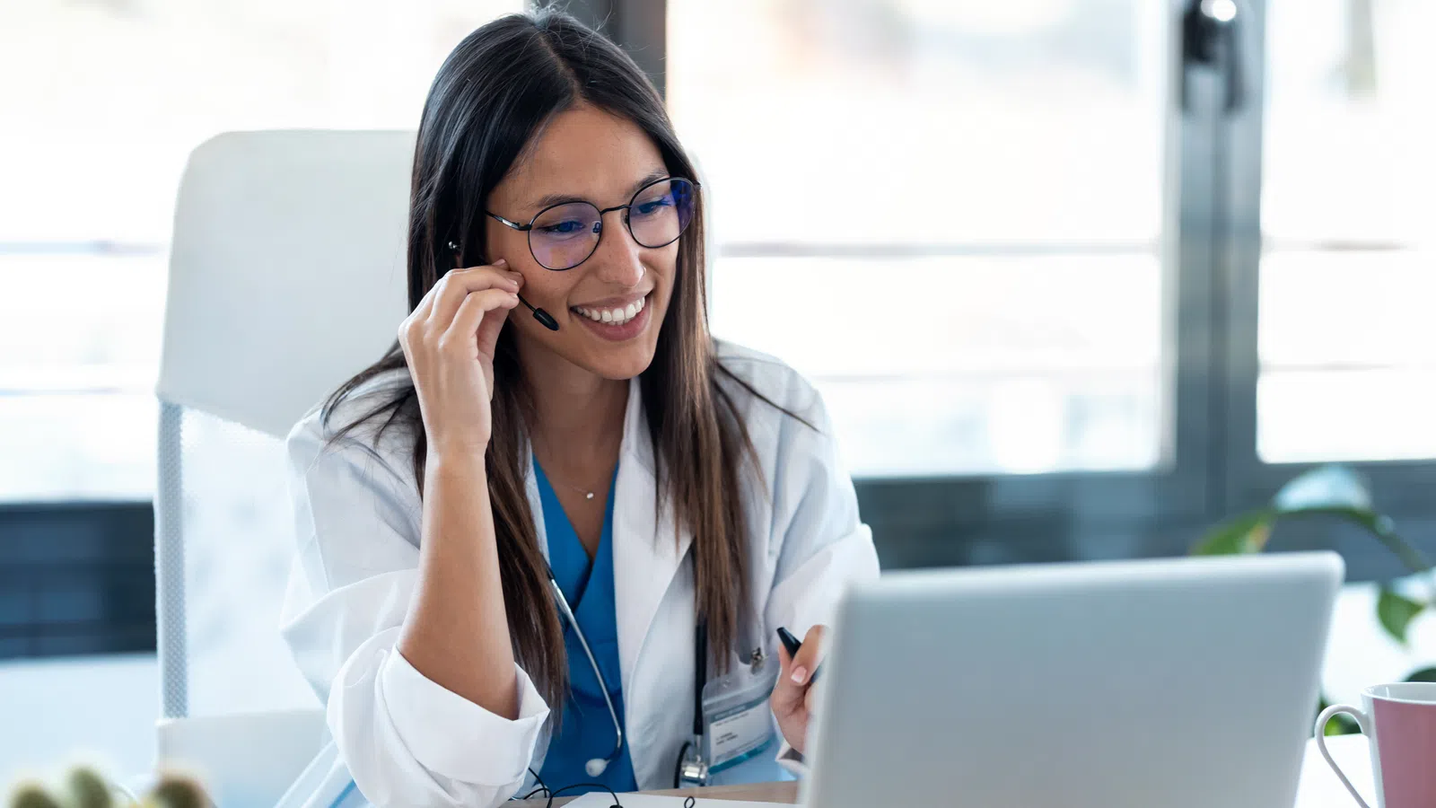 Female doctor wearing headset and white coat smiling during online consultation on laptop at office desk.