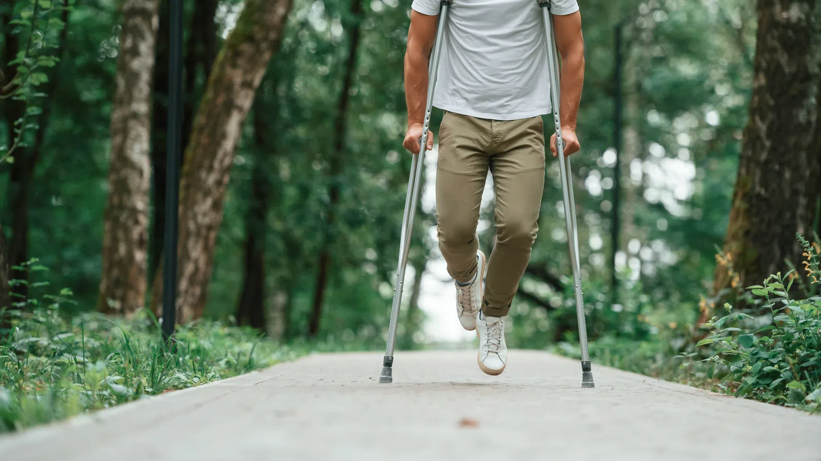 Man walking with crutches on a forest path surrounded by greenery, wearing a white shirt and beige pants.