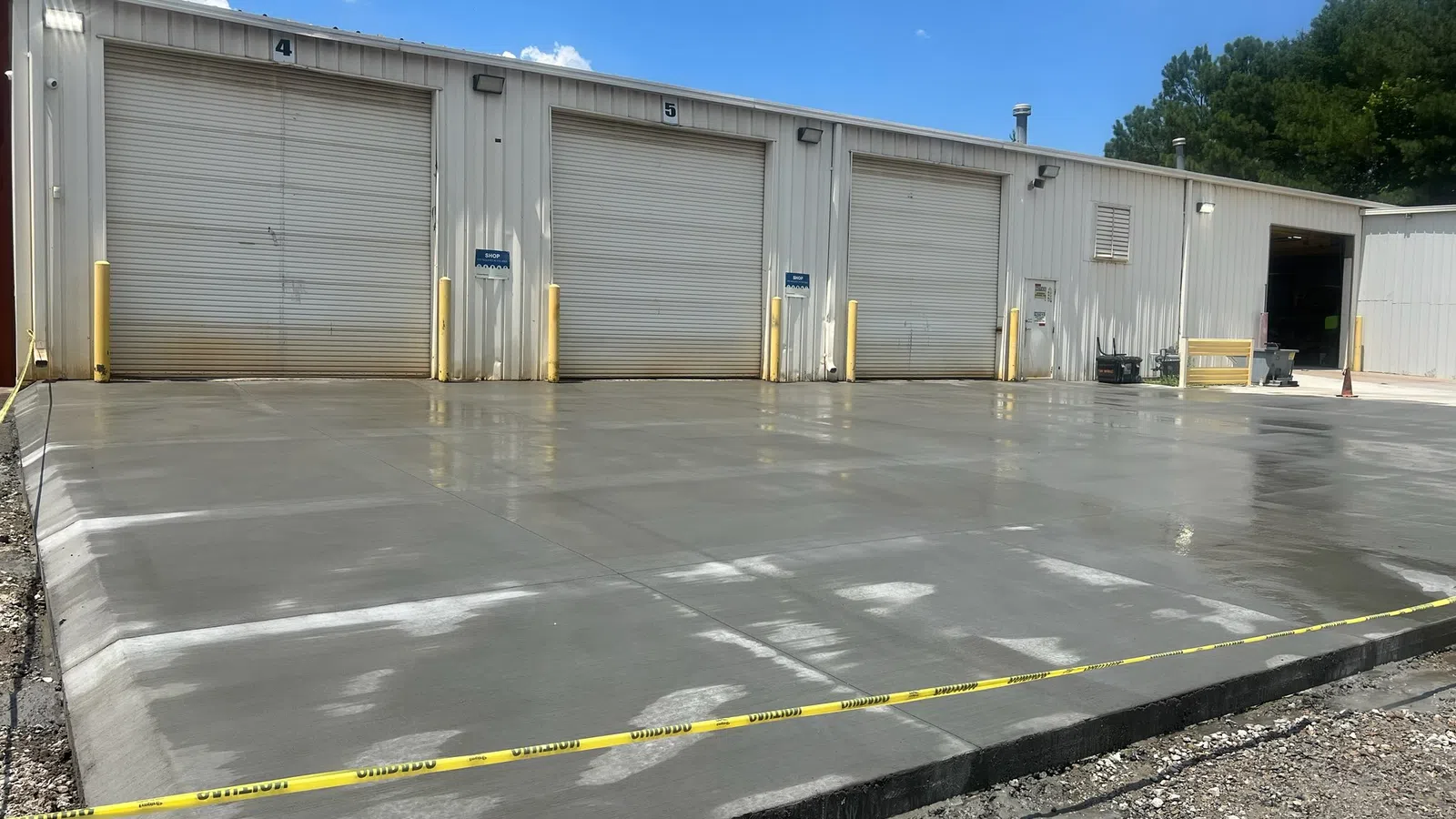 Newly poured concrete slab in front of warehouse garage doors with caution tape around the area under a blue sky.