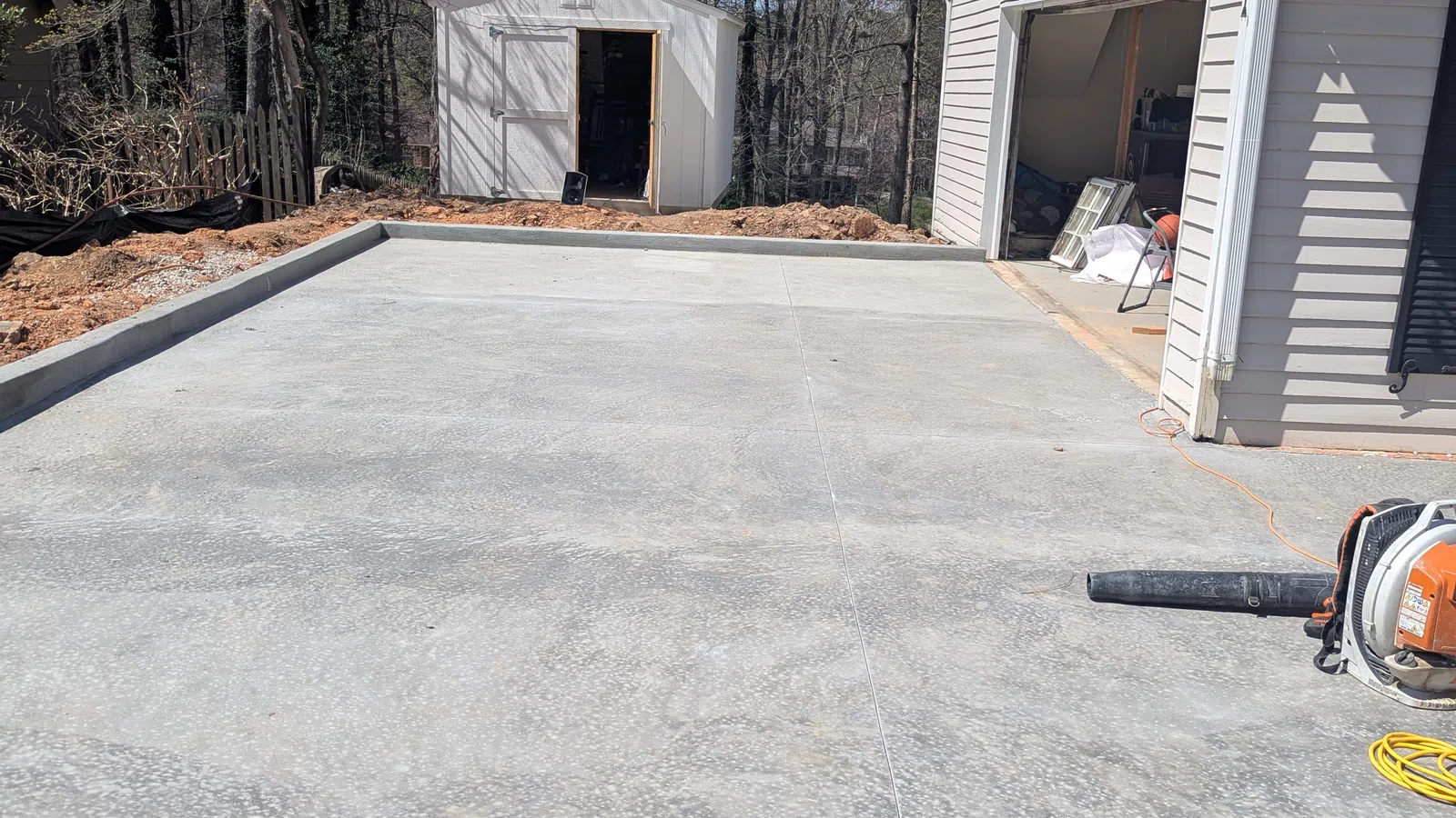 Freshly poured concrete driveway next to a white garage and a small white storage shed surrounded by trees.