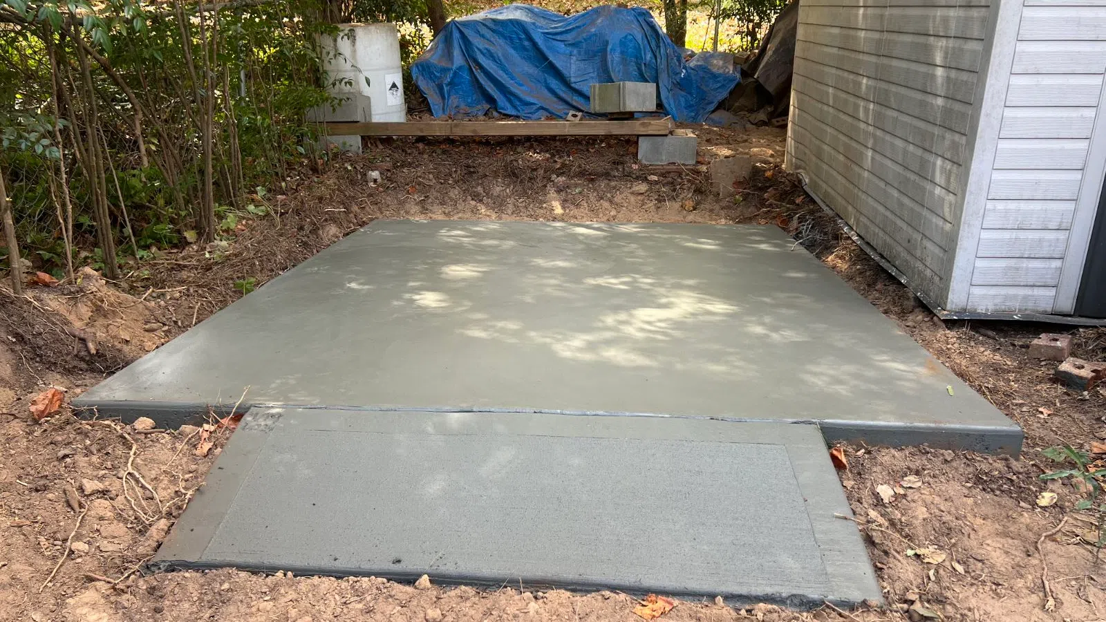 Newly poured smooth concrete slab next to a white shed in a backyard setting with trees and a blue tarp.