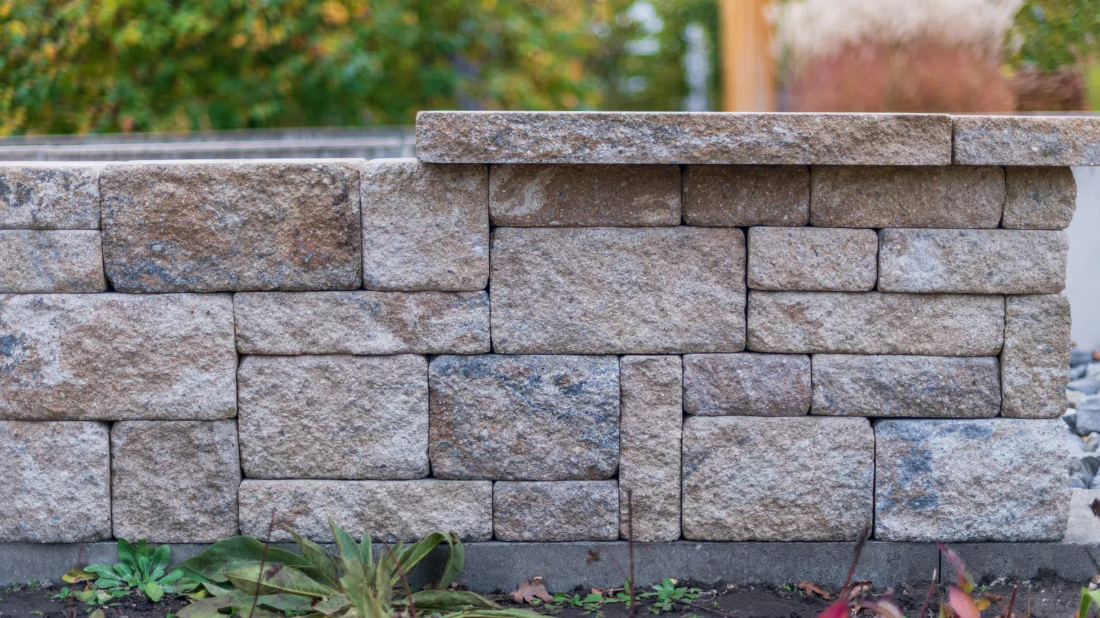 Close-up of a beige stacked stone retaining wall with garden plants and soil in the foreground.