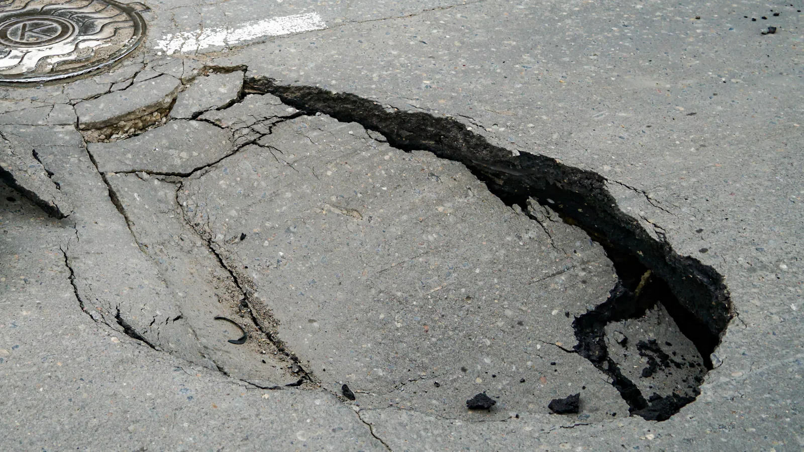 Cracked pavement with a large pothole and visible road markings, highlighting urban infrastructure issues.