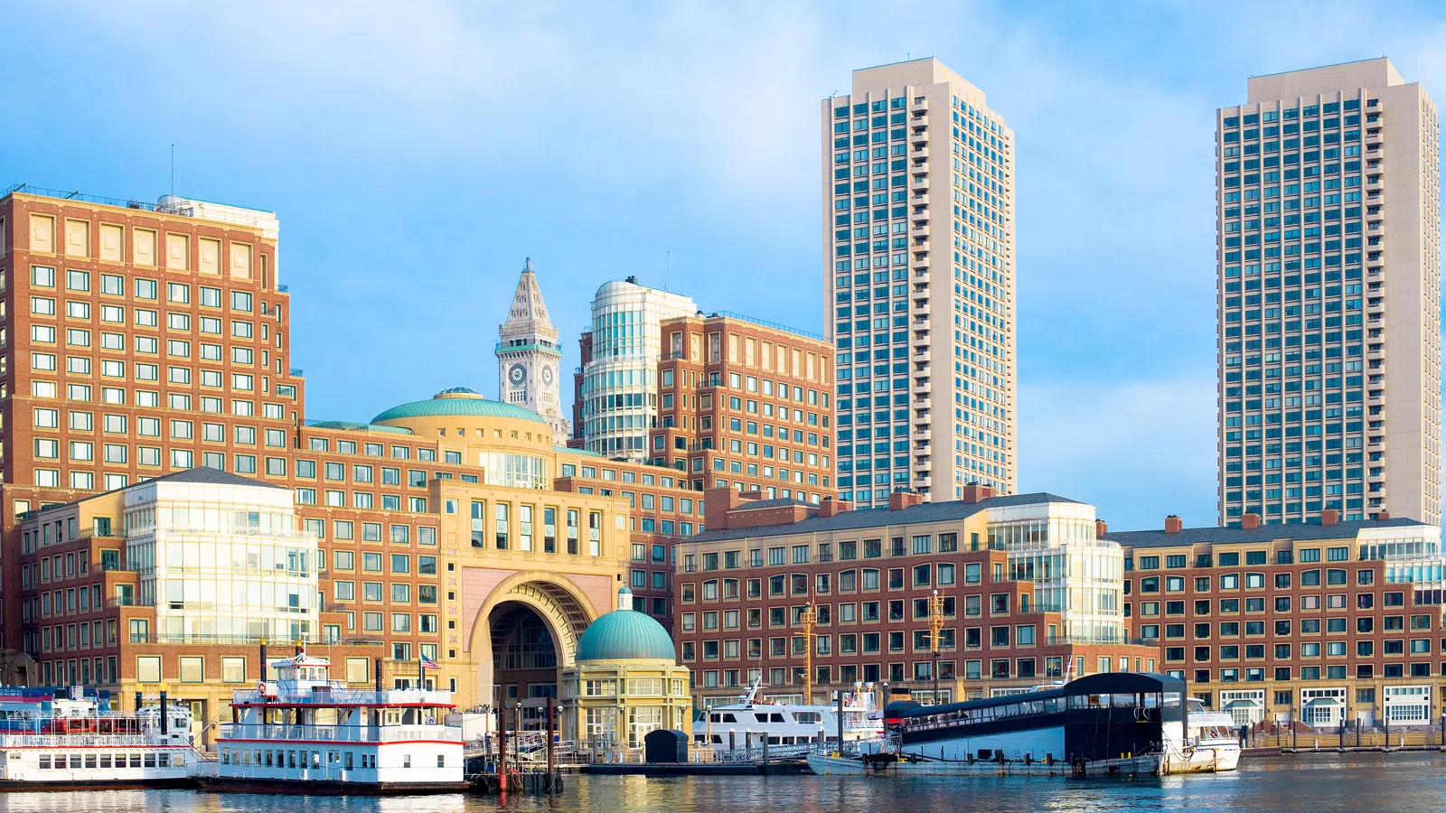 Boston waterfront with historic and modern buildings reflecting in calm harbor waters under blue sky