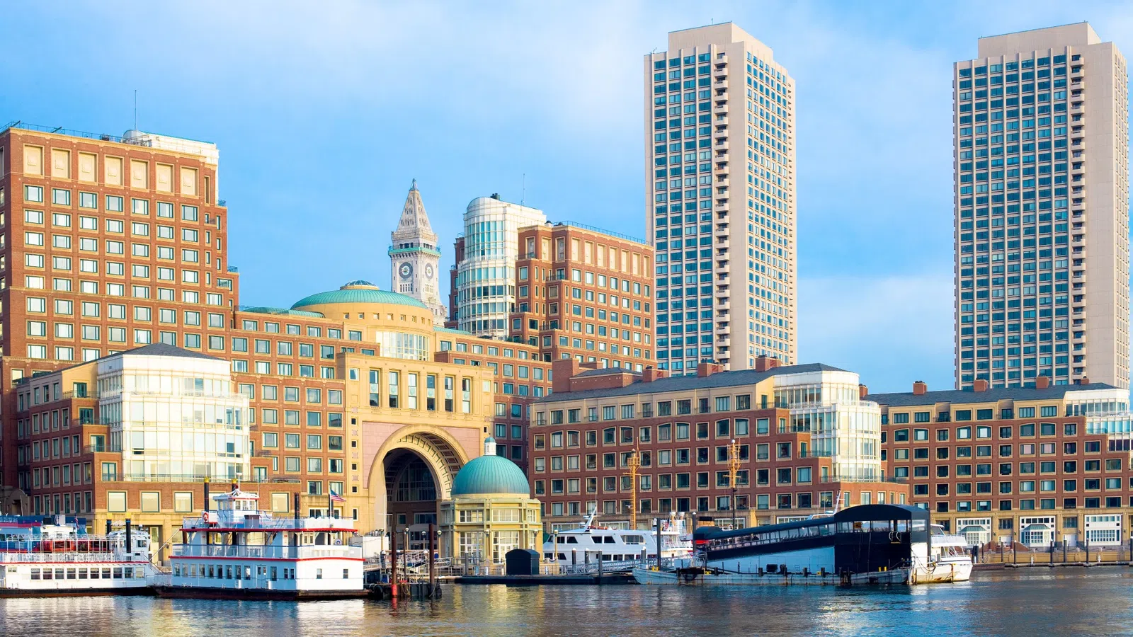 Waterfront cityscape with boats docked in calm harbor and modern buildings under clear blue sky.
