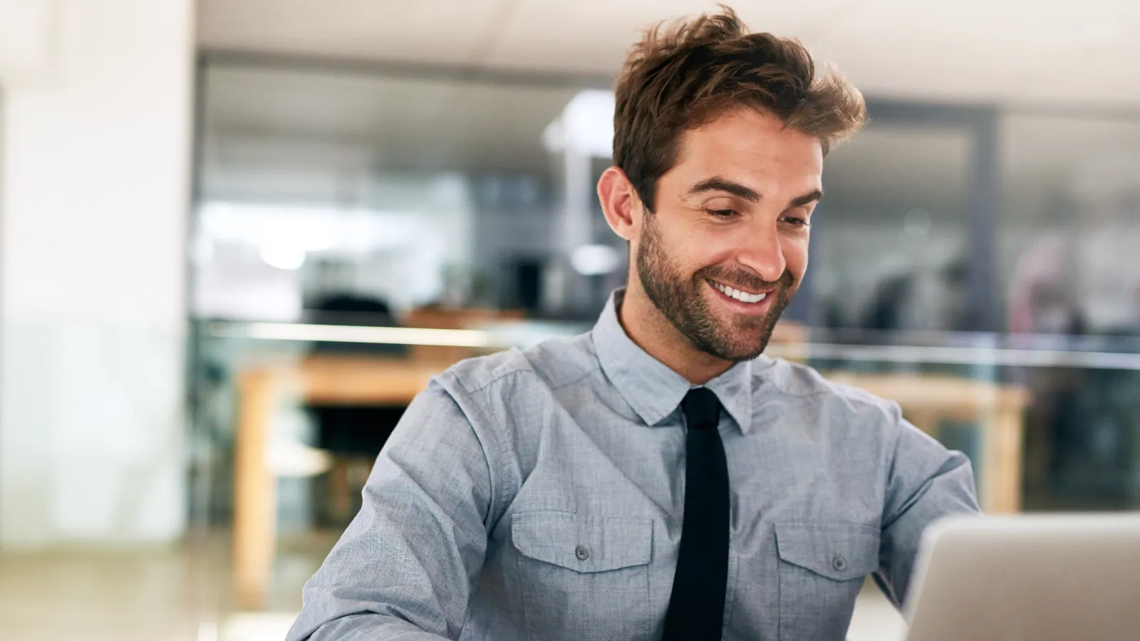 Smiling young businessman in gray shirt and black tie working on a laptop at office desk.