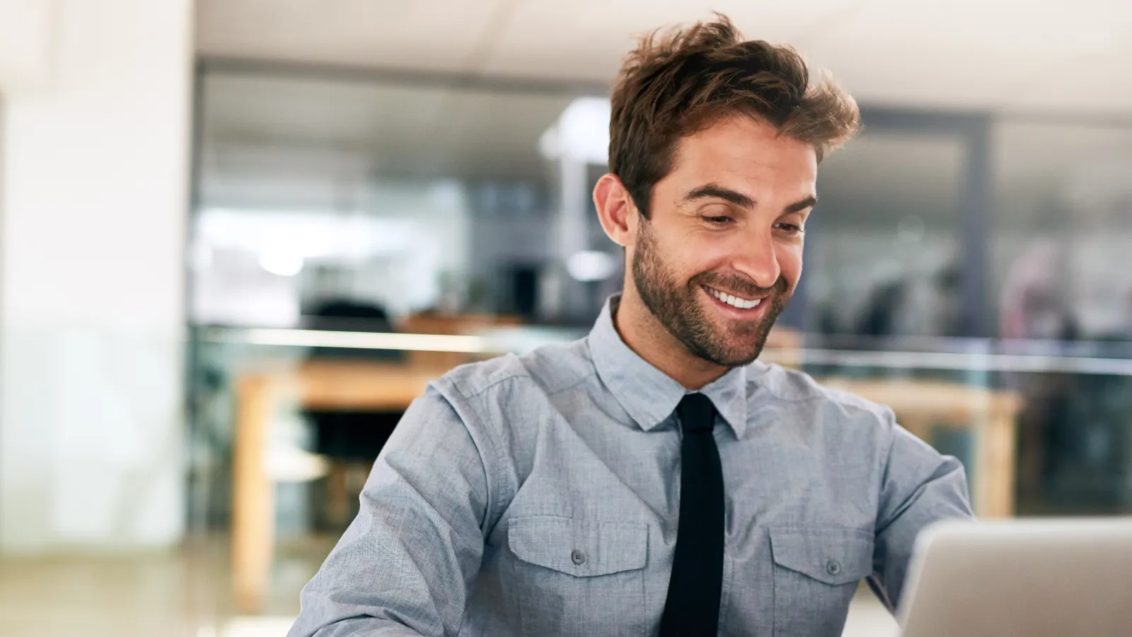 Smiling young man in shirt and tie working on a laptop at a wooden desk in a modern office setting