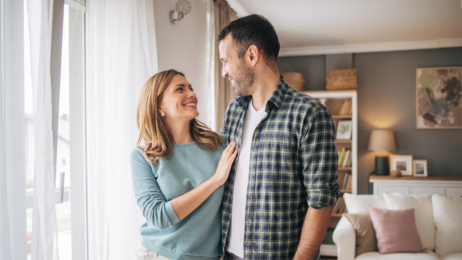 Couple smiling and embracing in a cozy, well-lit living room with modern decor and natural light.