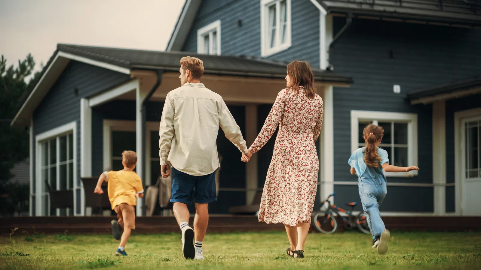 Family walking hand in hand toward a modern blue house with children running on the grass yard.