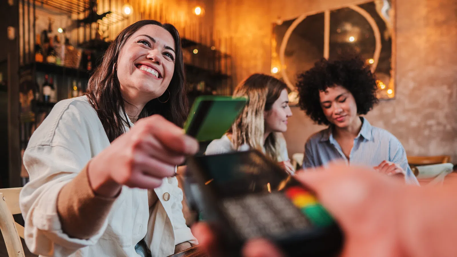 Smiling woman paying with credit card at cafe terminal while friends chat in background