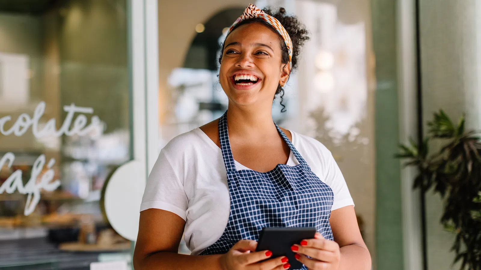 Smiling barista wearing apron and headband holds mobile phone inside modern cafe with glass windows.
