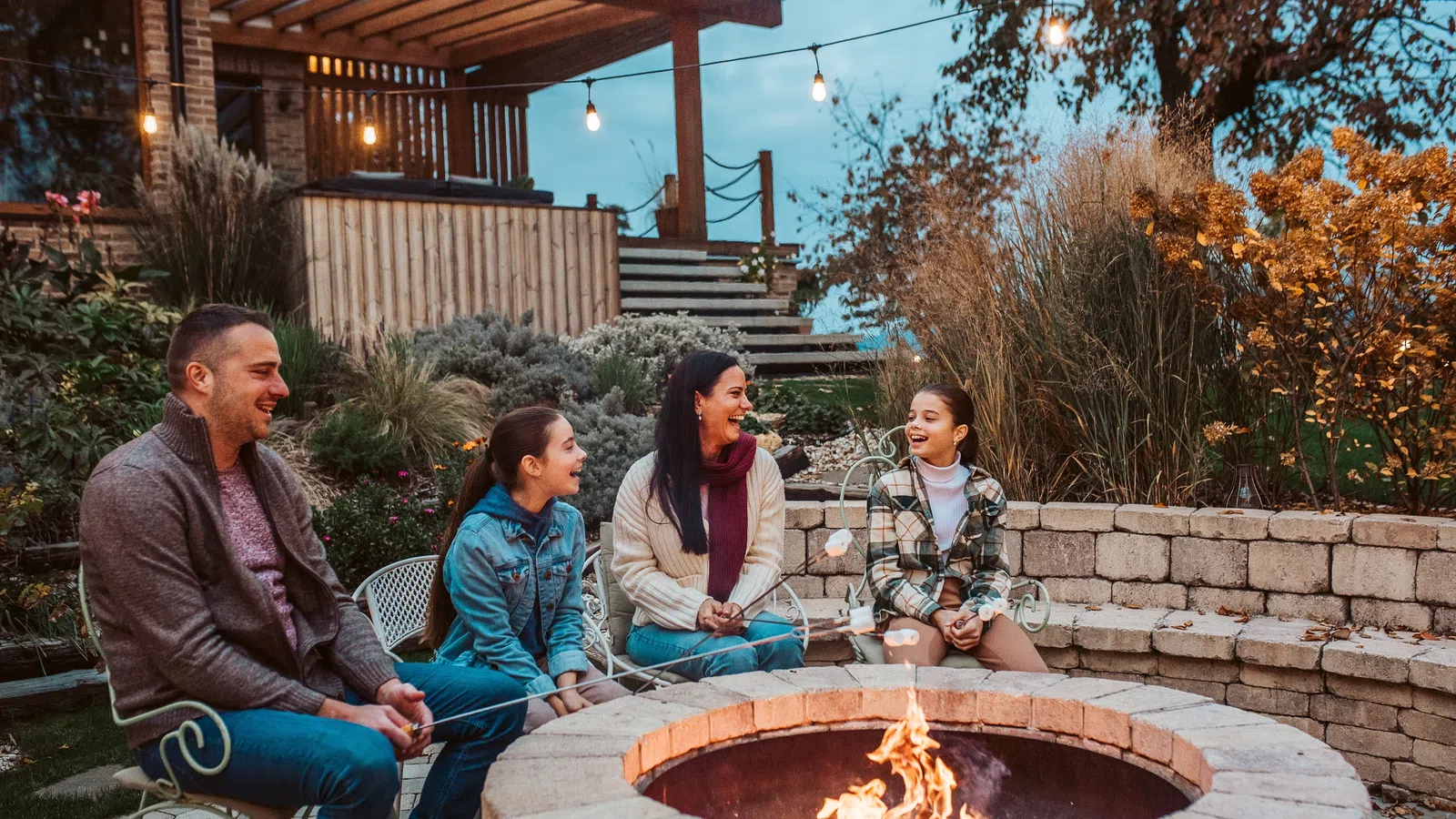 Family roasting marshmallows around a stone fire pit on a cozy evening outdoors with warm lights and fall foliage.