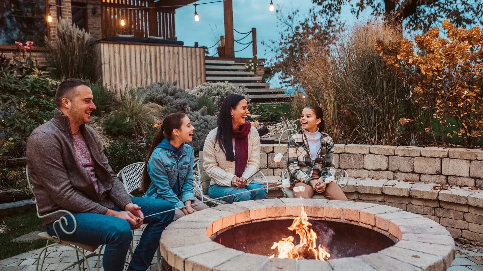 Family roasting marshmallows around a stone fire pit on a cozy evening outdoors with warm lights and fall foliage.