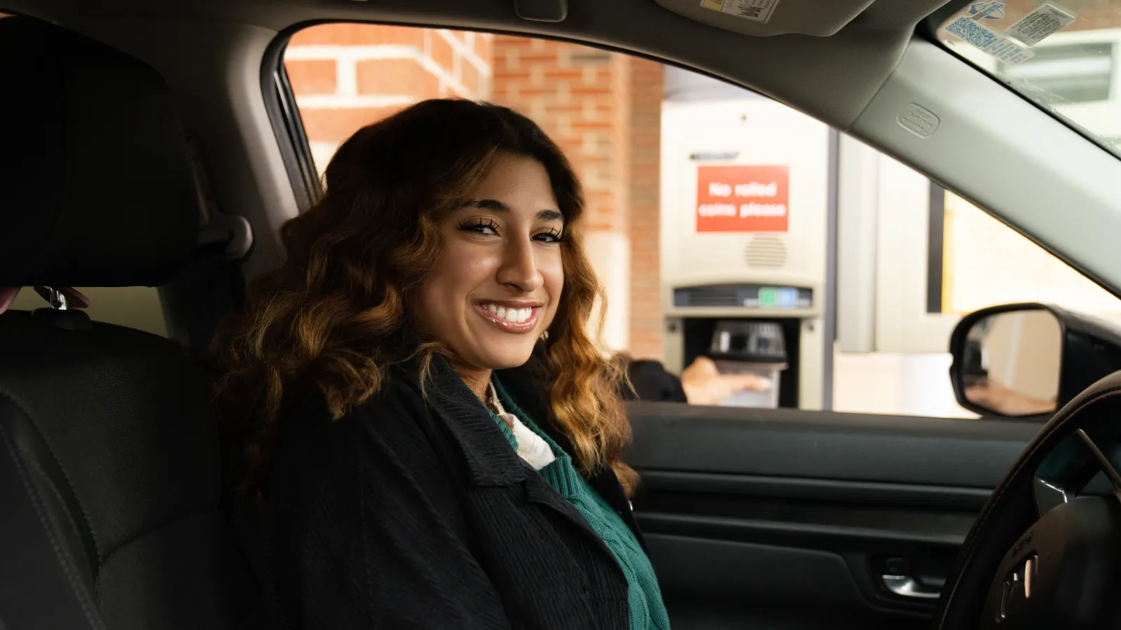 Happy woman sitting in car using a drive-thru ATM machine with brick wall background