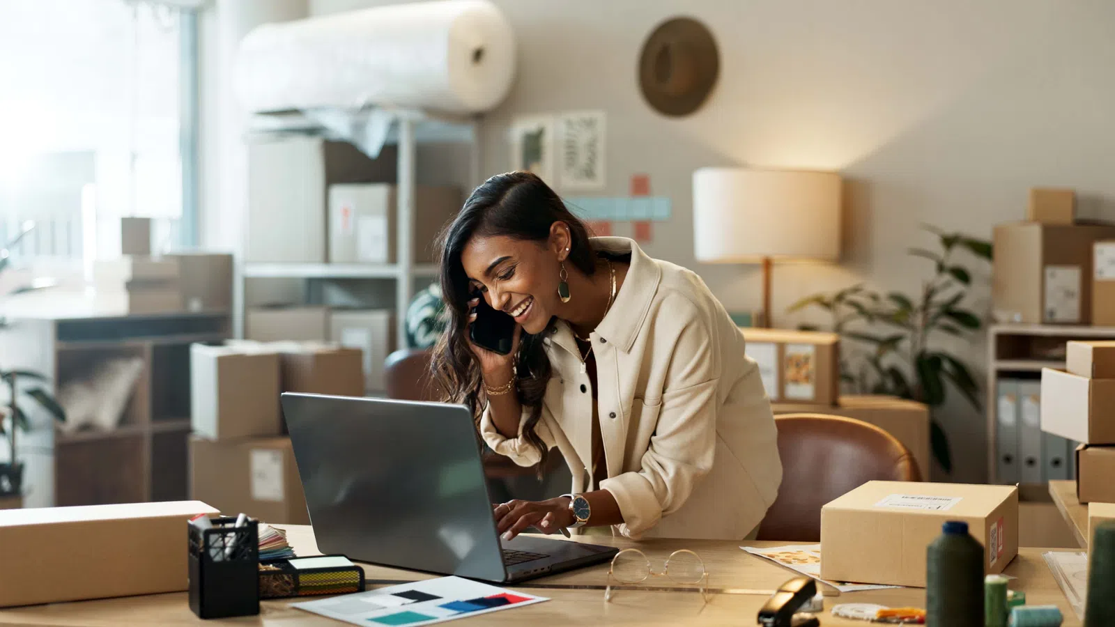 Woman smiling, talking on phone, and working on laptop surrounded by shipping boxes in a home office.