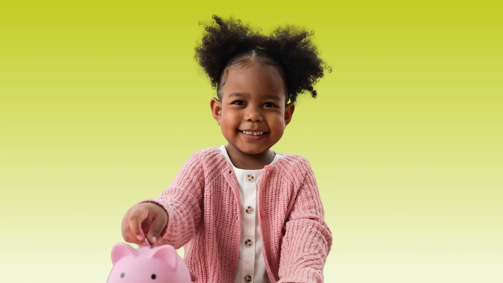 Smiling young girl in pink sweater putting coin into pink piggy bank on wooden table