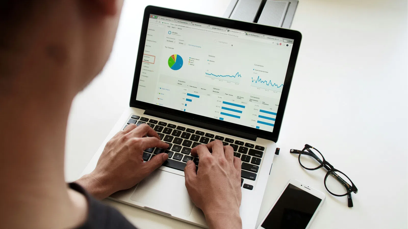 Person analyzing business analytics on a laptop with charts next to smartphone and eyeglasses on white desk.