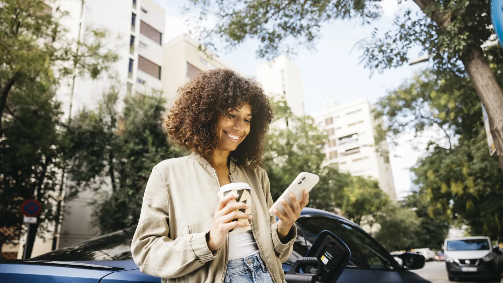 Young woman holding coffee and smartphone while charging electric car outdoors in city street.