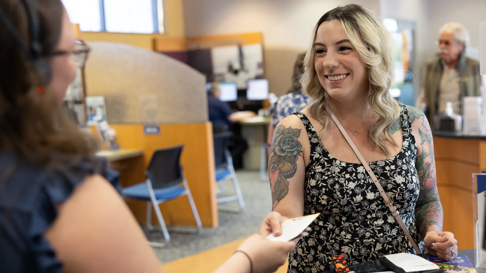Smiling young woman with tattoos handing a card to a receptionist in a busy office environment.