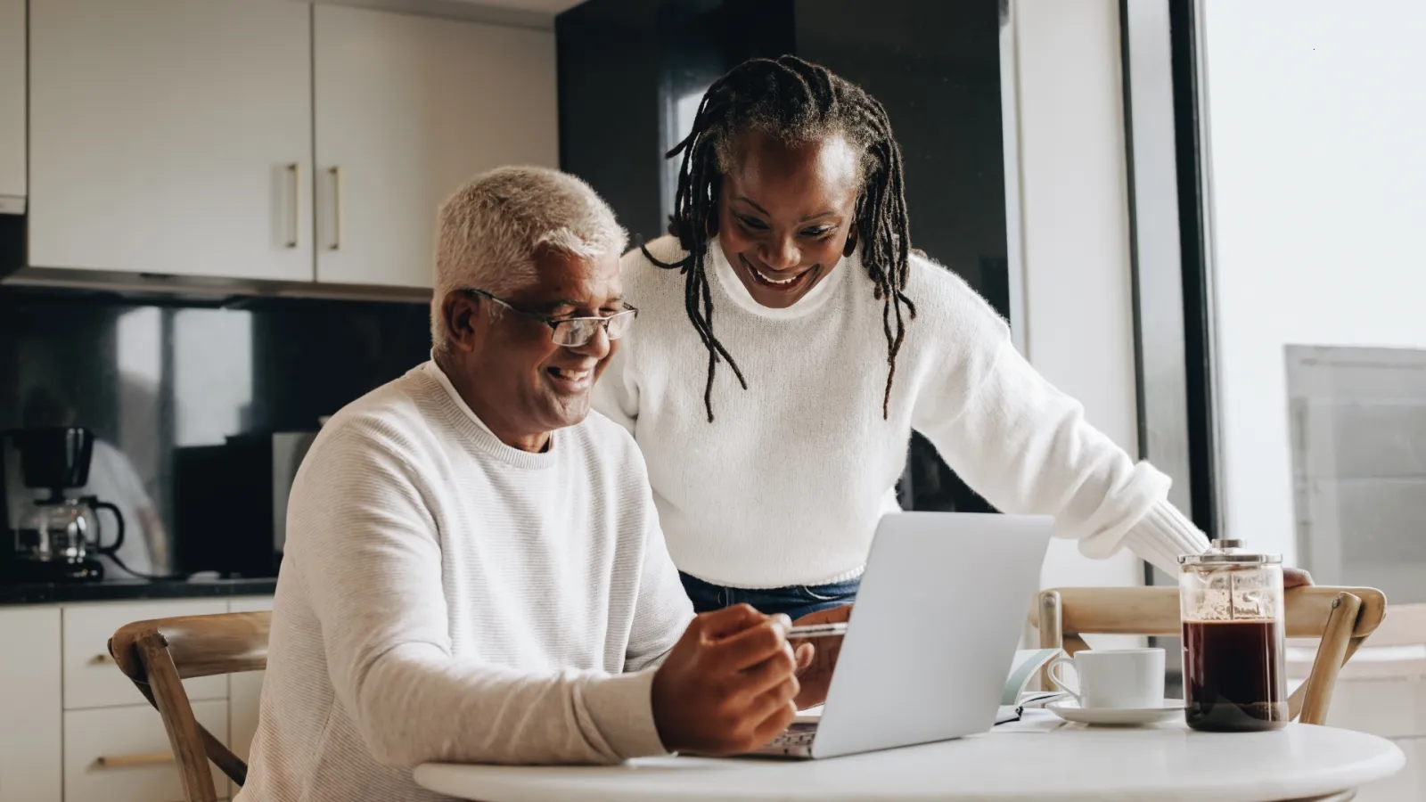 Smiling mature man and young woman using laptop together at kitchen table in bright modern home.