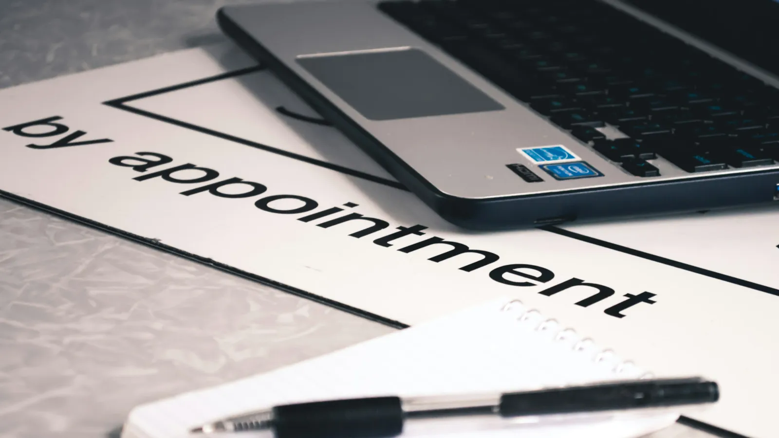 Laptop on a desk with a note indicating 'by appointment' and a pen resting on a notebook.