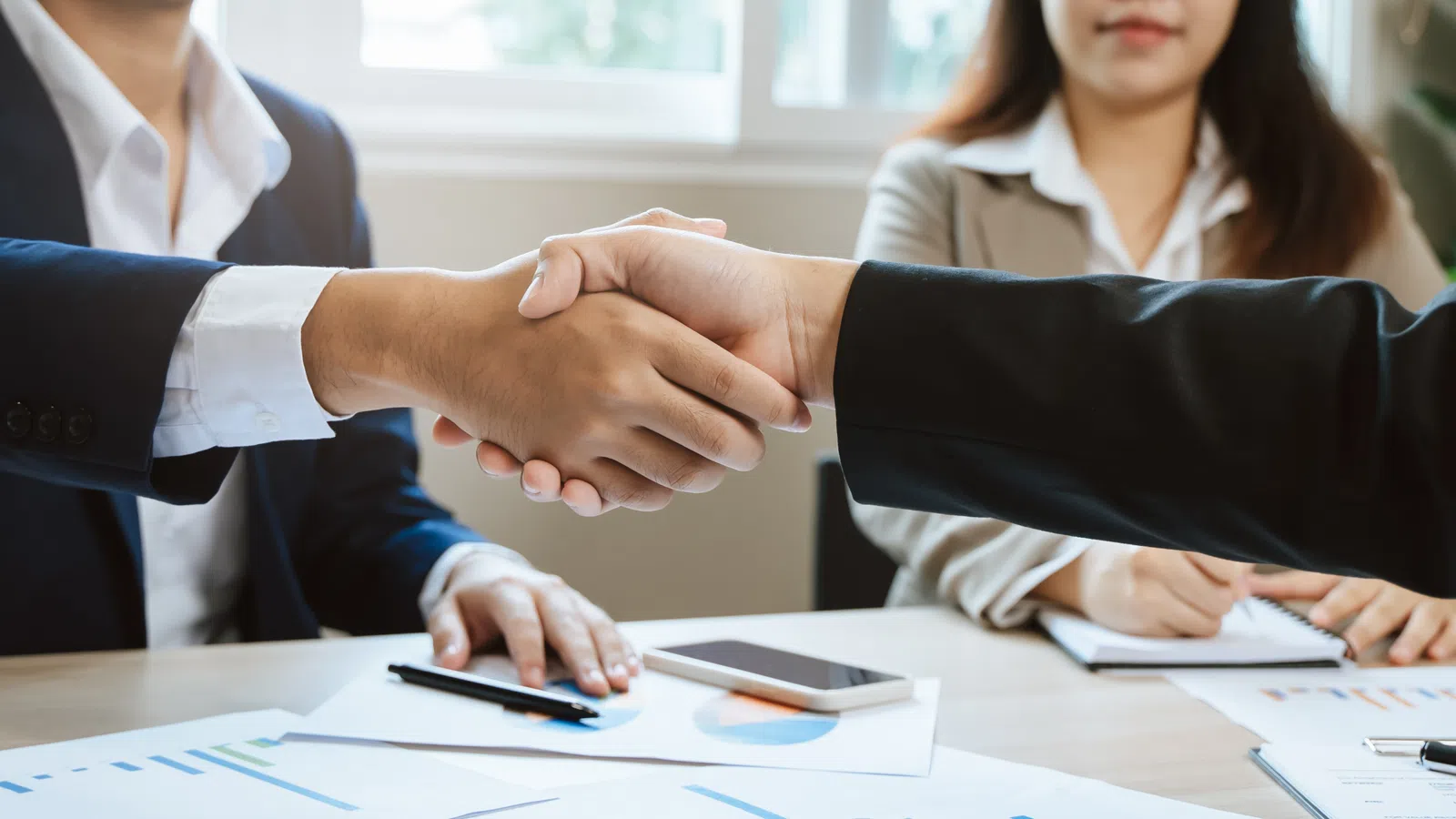 Two business professionals shaking hands over a table with documents and smartphone, signifying agreement.