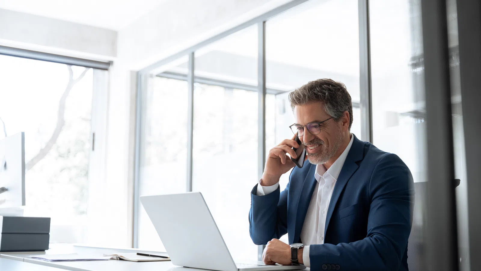 Man in blue suit smiling while talking on phone at desk with laptop in bright modern office
