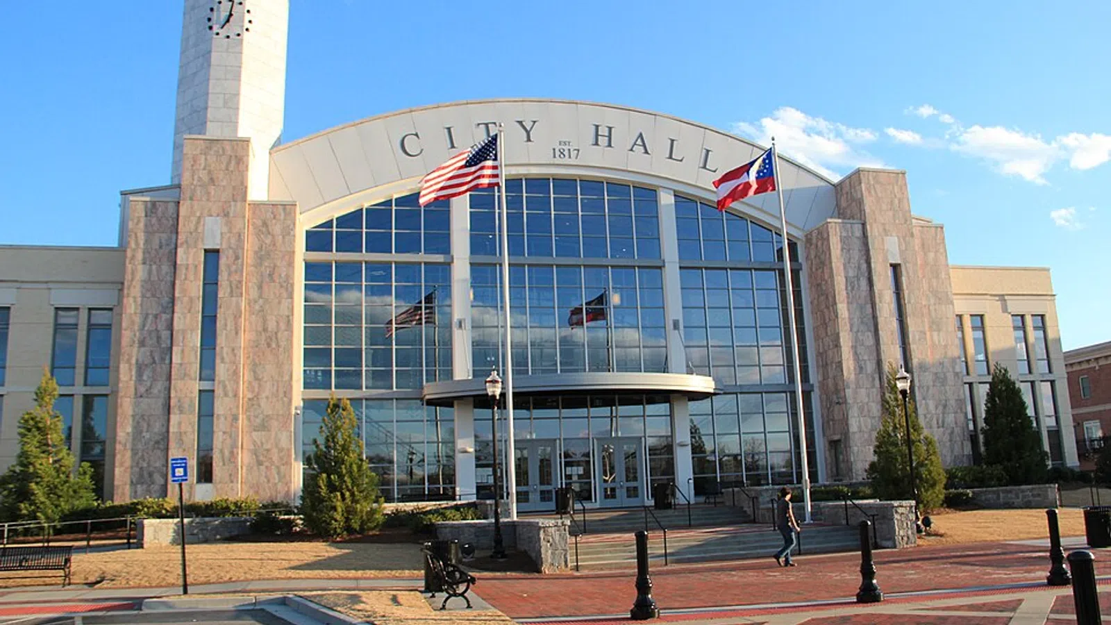 Modern city hall building with large glass windows, flags, clock tower, and clear blue sky in the background.