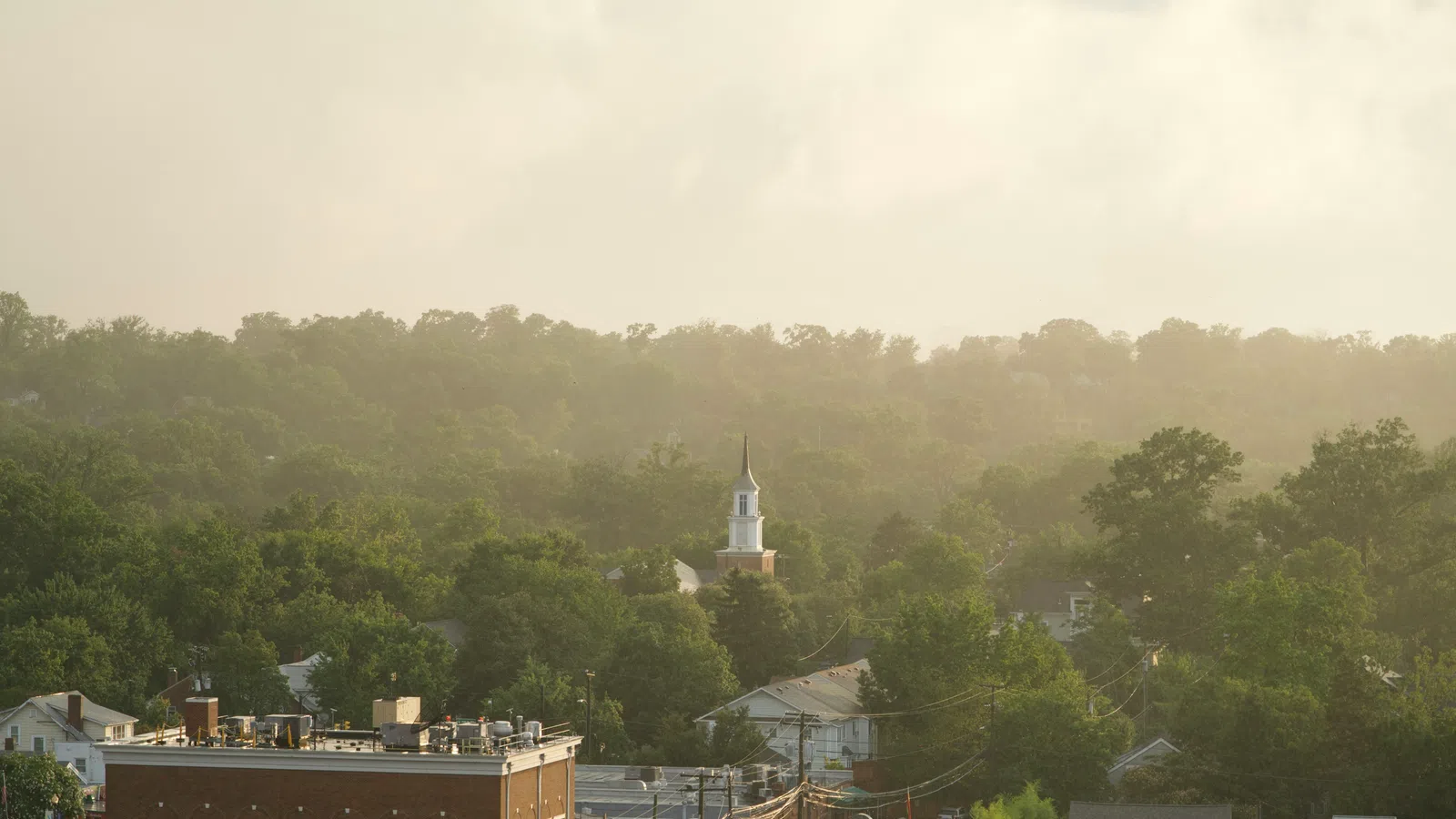 Small town with dense green trees, houses, a church steeple, and hazy sunlight in the background.