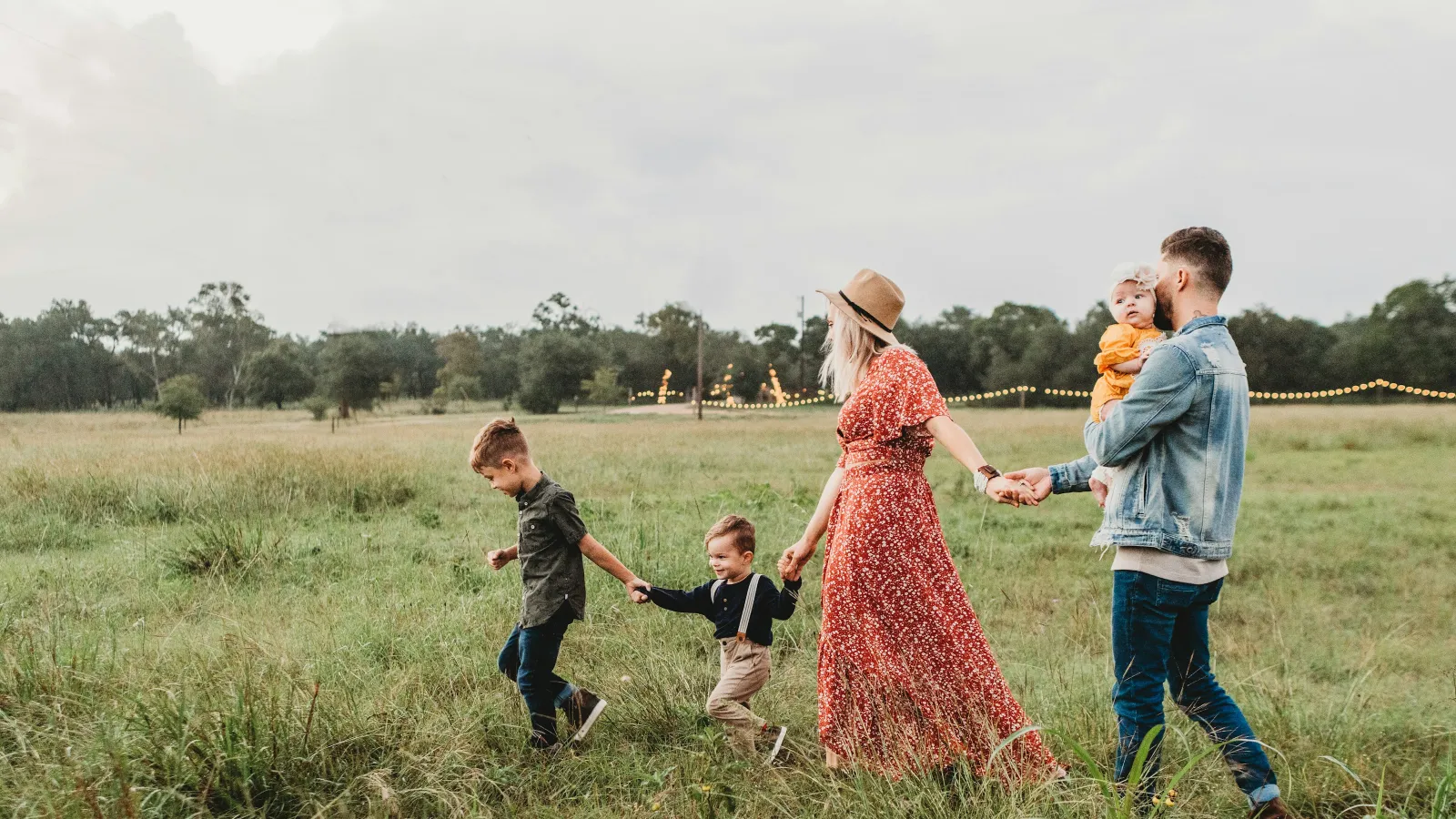 Family of five walking hand-in-hand through a grassy field with string lights in the background during cloudy day