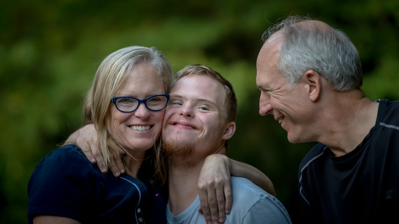 Smiling family with a young man with Down syndrome embracing outdoors with green background.