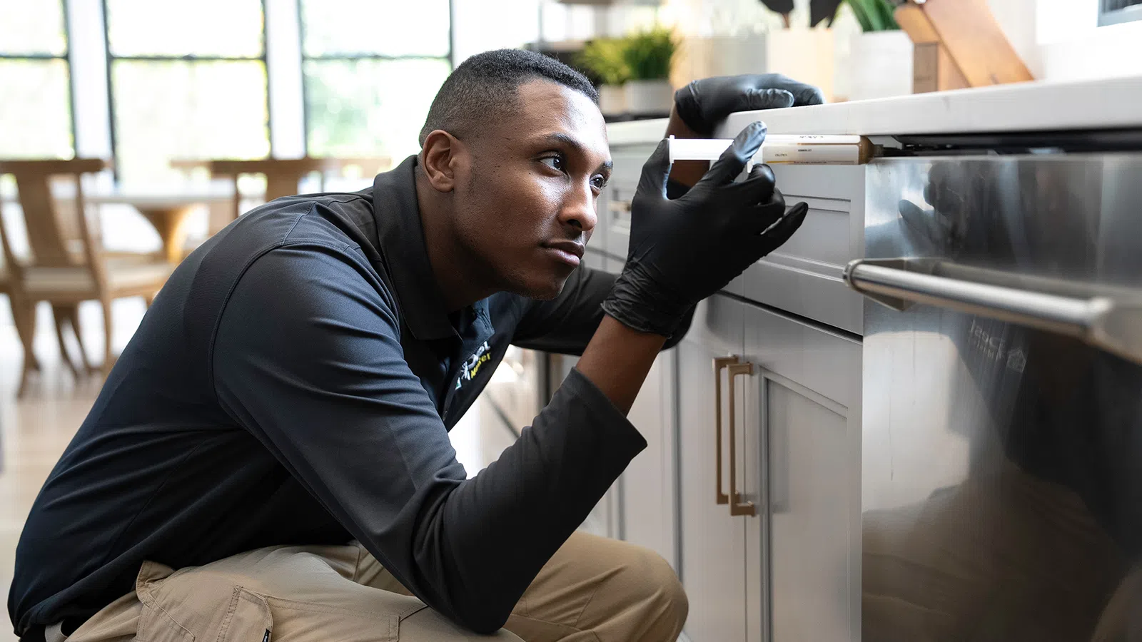 Technician in black gloves inspecting kitchen appliances for pests using a detection tool in a bright home.