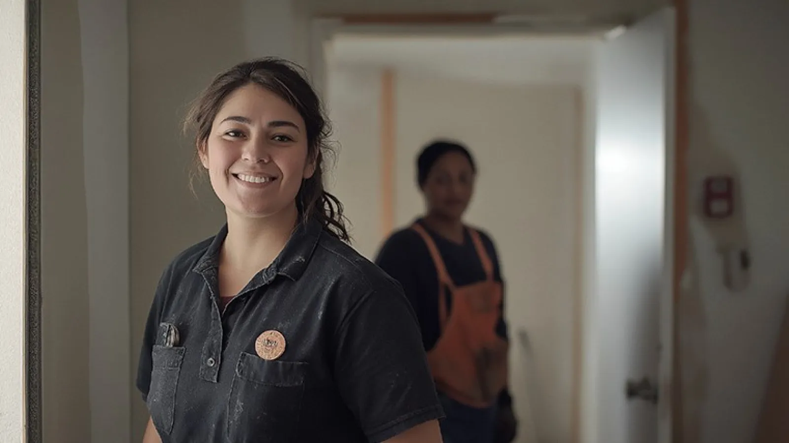 Smiling female construction worker in casual workwear stands indoors with another worker in background.