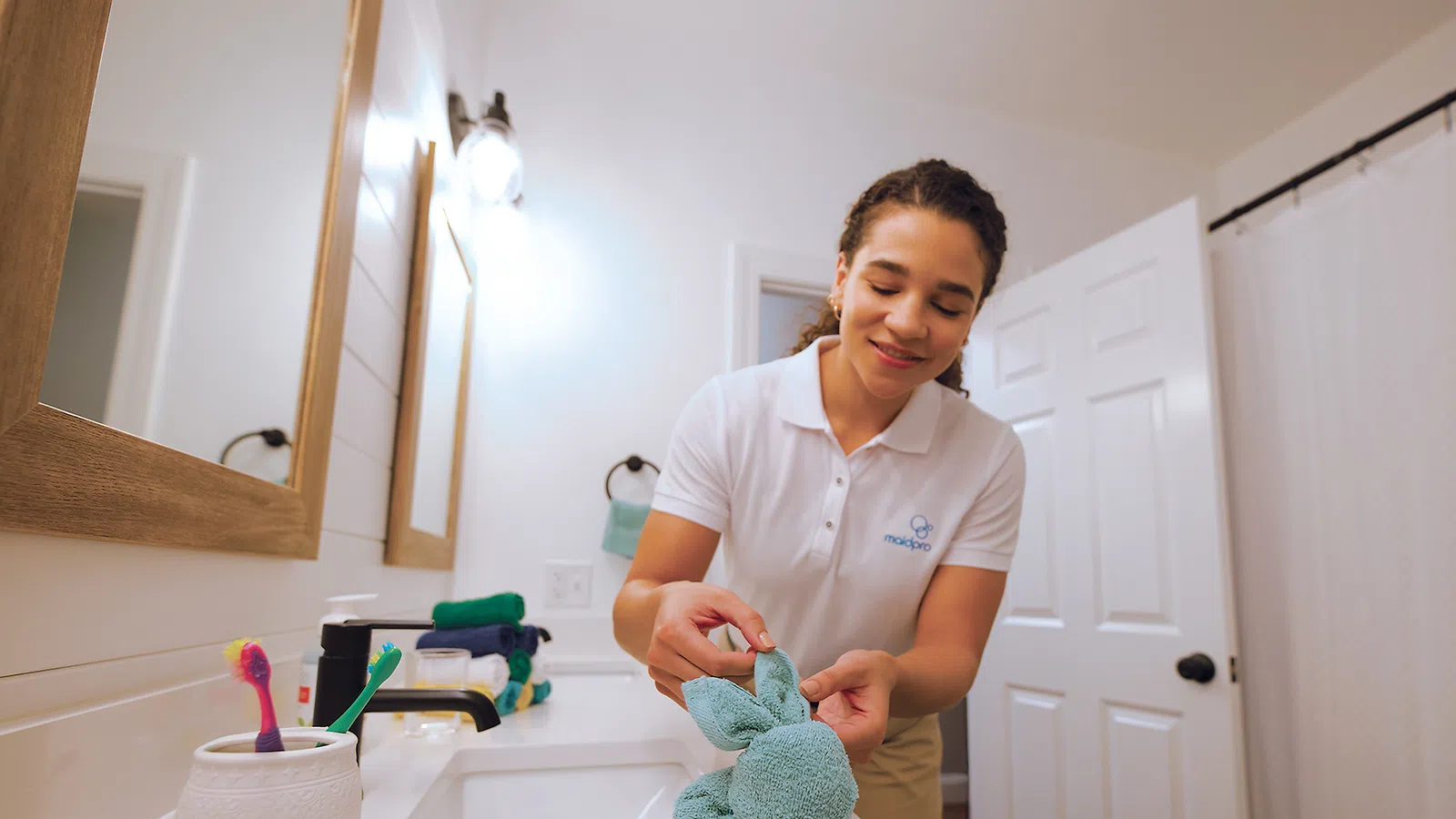 Woman in uniform shaping a green towel into a bunny on a bathroom counter with toothbrushes and mirror.