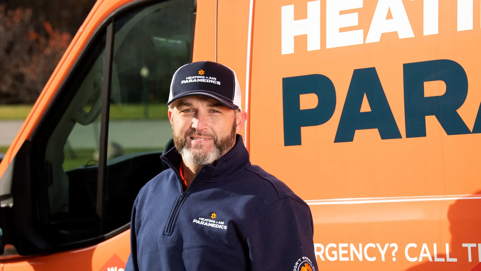 Smiling technician in uniform stands beside orange Heating & Air Paramedics service van outdoors.
