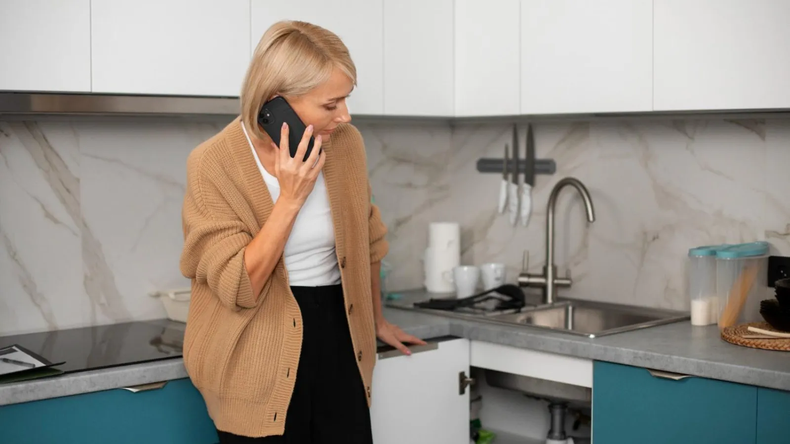 Woman in brown cardigan talking on phone while inspecting kitchen cabinet under sink in modern kitchen.