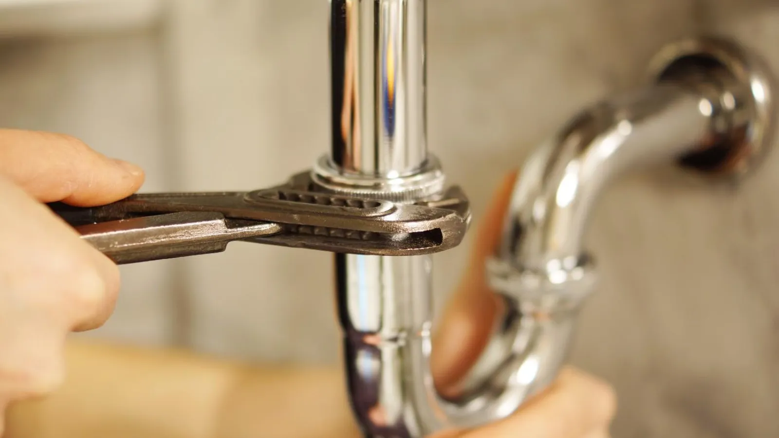 Hand tightening a chrome pipe under a sink with an adjustable wrench during plumbing repair.