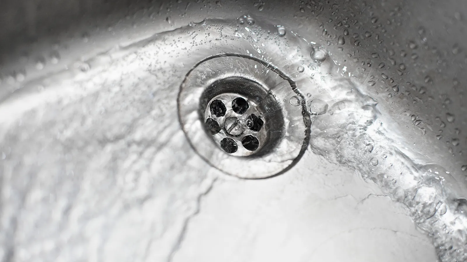 Close-up of water flowing down a stainless steel sink drain with water droplets on the surface