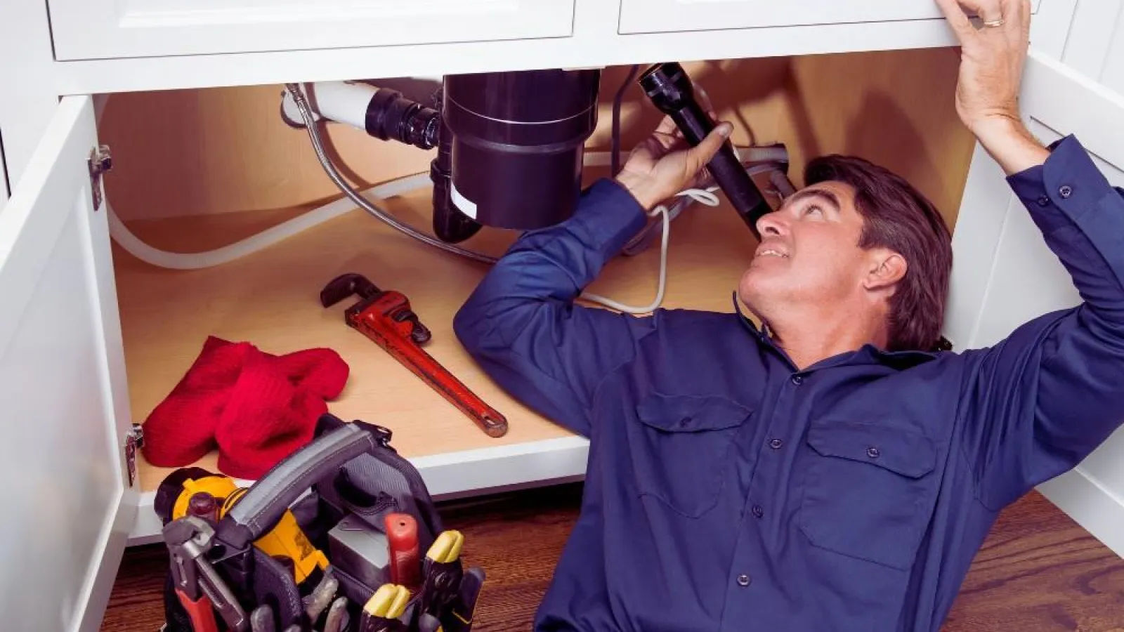 Plumber in blue uniform lying on wooden floor fixing pipes under a white kitchen sink with flashlight and tools