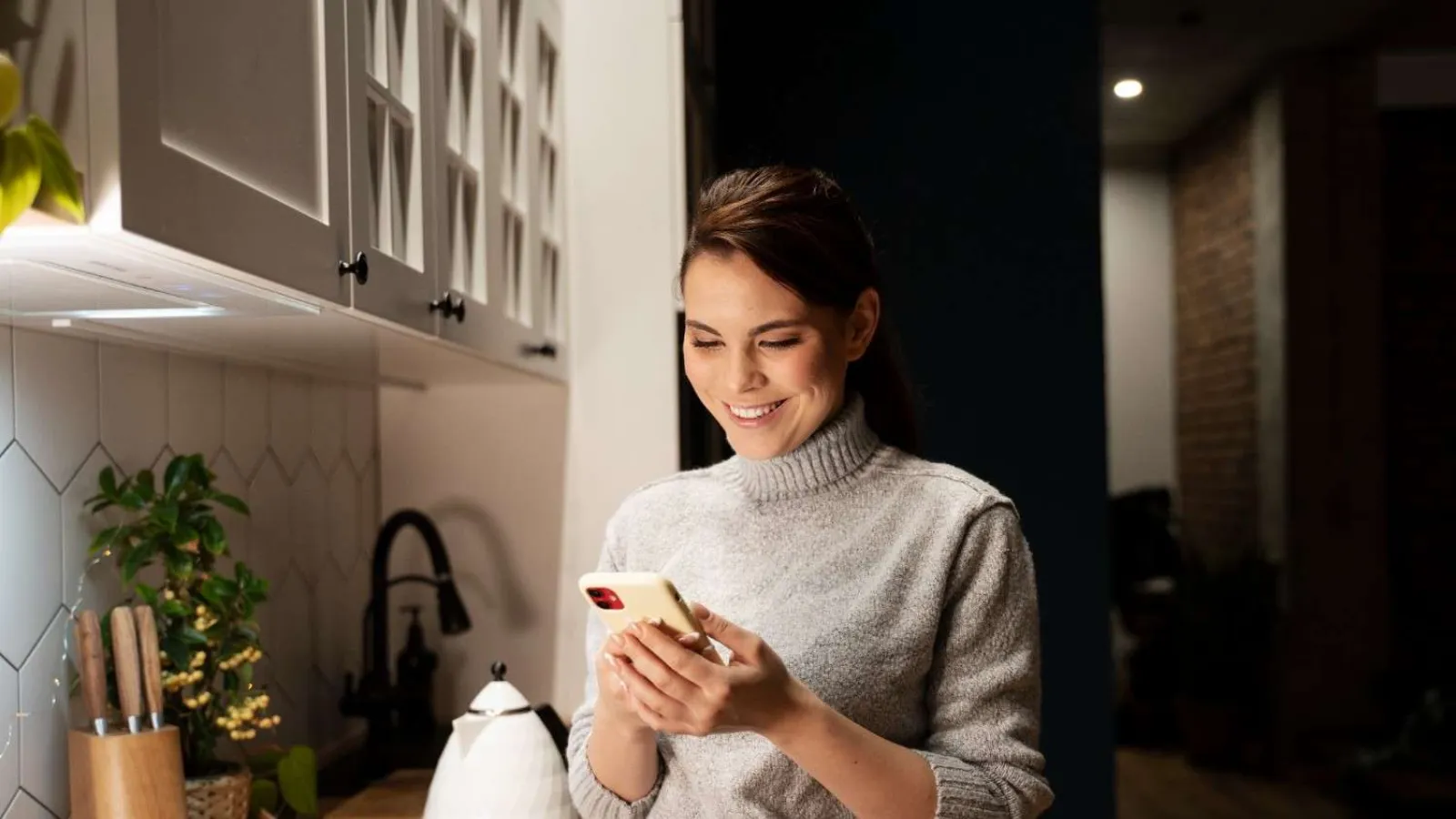 Woman in gray sweater smiling while using smartphone in a modern kitchen with wooden countertop.