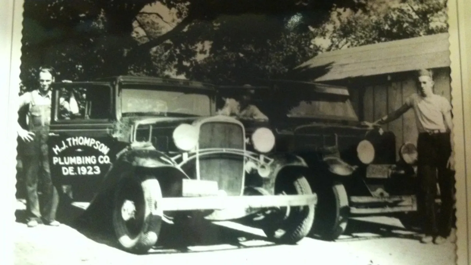 Black and white photo of two men standing beside vintage cars, one marked H.J. Thompson Plumbing Co. 1923.